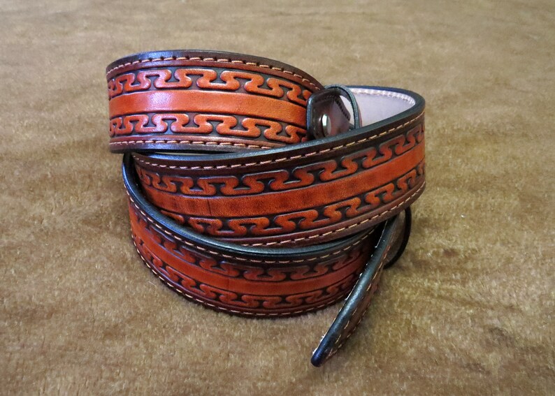 May include: A brown leather belt with a decorative pattern of interlocked lines. The belt is folded in half and the edges are stitched with a dark thread.