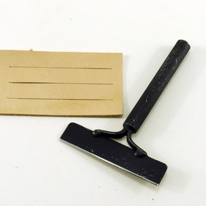 May include: A black leather working tool with a handle and a blade. The tool is used to score leather for stitching. A piece of brown leather with lines scored into it is also shown.