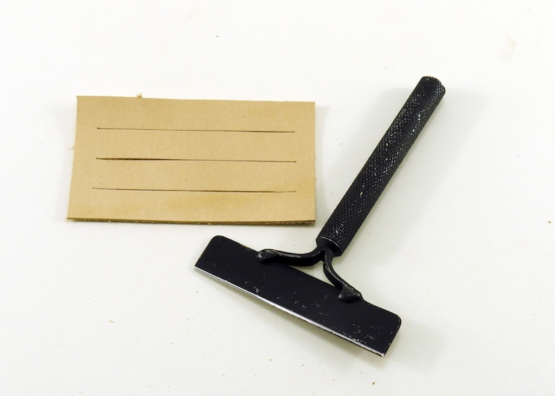 May include: A black leather working tool with a handle and a blade. The tool is used to score leather for stitching. A piece of brown leather with lines scored into it is also shown.