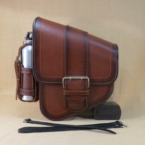 Può includere: Sella da moto in pelle marrone con chiusura a fibbia e una tasca laterale per una bottiglia d'acqua. La borsa è fissata a una cinghia nera.