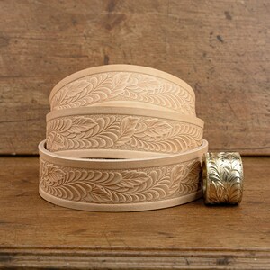 May include: Three stacked leather belts with a floral design. The belts are a light brown color and have a smooth texture. A gold metal belt buckle is next to the belts.