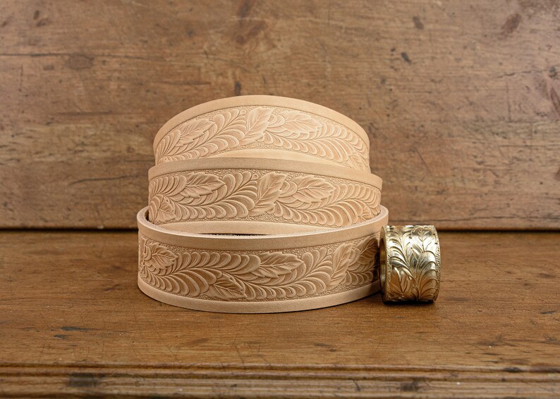 May include: Three stacked leather belts with a floral design. The belts are a light brown color and have a smooth texture. A gold metal belt buckle is next to the belts.