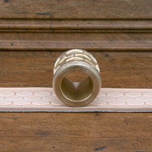 May include: A gold-colored metal roller with a hexagonal pattern on the surface. The roller is resting on a light pink leather strap with a hexagonal pattern.