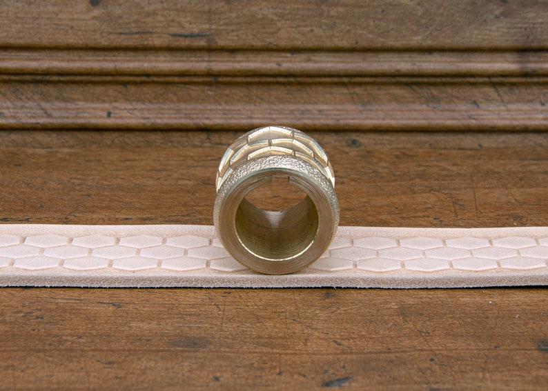 May include: A gold-colored metal roller with a hexagonal pattern on the surface. The roller is resting on a light pink leather strap with a hexagonal pattern.