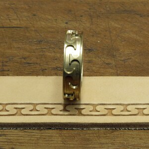 May include: A gold-colored leather stamping tool with a repeating pattern of interlocking S shapes. The tool is being used to create a border on a piece of leather.