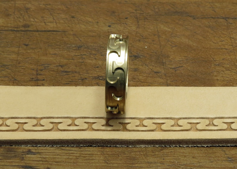 May include: A gold-colored leather stamping tool with a repeating pattern of interlocking S shapes. The tool is being used to create a border on a piece of leather.
