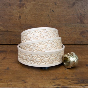 May include: Three leather belts with an intricate woven design, stacked on top of each other. The belts are a light cream color and have a smooth, polished finish. A small, gold-colored metal buckle is also visible in the image.
