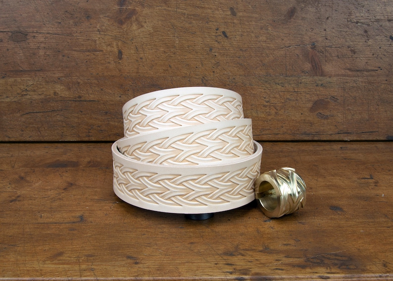 May include: Three leather belts with an intricate woven design, stacked on top of each other. The belts are a light cream color and have a smooth, polished finish. A small, gold-colored metal buckle is also visible in the image.