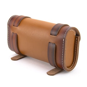 May include: A brown leather cylindrical case with decorative stitching and straps. The case has a flap that closes with a snap.