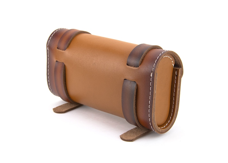 May include: A brown leather cylindrical case with decorative stitching and straps. The case has a flap that closes with a snap.