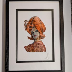 Puede incluir: Obra de arte enmarcada que presenta un retrato estilizado de una mujer con cabello naranja y lápiz labial rojo. La palabra "MONSTER" es visible en el cabello. La obra de arte está en un marco negro y está firmada en la esquina inferior derecha.