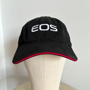 May include: Black baseball cap with the word "EOS" embroidered in white on the front. The cap has a red trim along the brim and is displayed on a mannequin head.