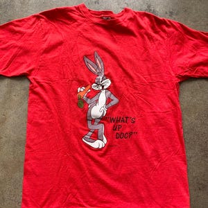 May include: Red t-shirt featuring Bugs Bunny holding a carrot and saying "What's Up Doc?"