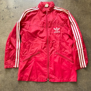 Op de afbeelding: Een rode Adidas-trainingsjas met witte strepen op de mouwen en het Adidas-logo op de borst. De jas heeft een volledige ritssluiting, twee voorzakken en een opstaande kraag. De jas is gemaakt van een lichtgewicht materiaal.