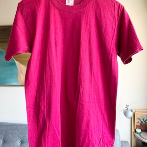 May include: A solid magenta-colored t-shirt with a crew neck and short sleeves. The shirt is hanging on a wooden hanger. The fabric appears to be a soft, textured material. The shirt is displayed against a neutral background.