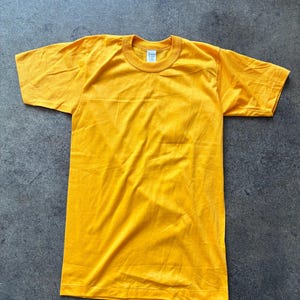 以下が含まれることがあります： 無地の黄色の半袖Tシャツ。Tシャツはクルーネックで、襟の内側にタグが付いています。生地は柔らかく、軽量に見えます。Tシャツは灰色の表面に平らに置かれています。