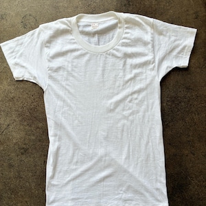 May include: A plain white t-shirt with a crew neck and short sleeves. The shirt is made of a soft, slightly textured fabric. A small tag is visible at the back of the neck. The shirt is laid flat on a brown surface.