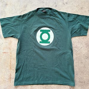 May include: A dark green t-shirt with the Green Lantern logo. The logo is a white circle with a green lantern symbol in the centre. The shirt is made of a soft material and has short sleeves.