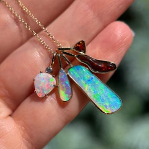 May include: A gold necklace featuring multiple gemstone pendants. The pendants include opals with iridescent colors, a heart-shaped opal, and a reddish-brown gemstone. The necklace is held in a hand.