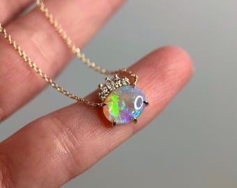 May include: A gold necklace featuring an oval opal pendant with a crown of small diamonds. The opal displays a vibrant play of color, including green, yellow, and blue hues. The chain is delicate, and the pendant is the focal point.