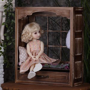 May include: A vintage-style wooden display cabinet with a doll seated inside. The doll has blonde hair and wears a plaid dress and white shoes. A small pillow and a decorative rug are also inside the cabinet.
