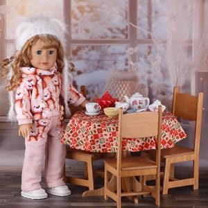 Puede incluir: Una muñeca de pelo largo y ondulado lleva un gorro blanco esponjoso y una sudadera con capucha con un patrón repetido de ositos de peluche y pantalones rosa claro a juego. La muñeca está junto a una pequeña mesa de madera con una tetera roja.