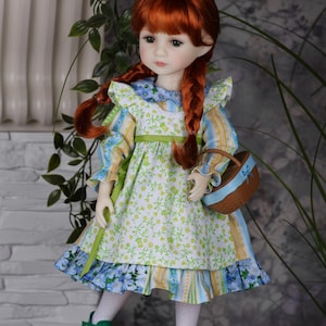 May include: A doll with red plaited hair, dressed in a floral dress with blue and yellow stripes, green shoes, and a small basket with a blue ribbon. The dress features a green ribbon belt and a blue floral skirt.