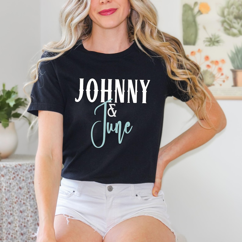 May include: Black t-shirt with the text "JOHNNY & June" in white and teal script. The shirt is a classic crew neck style. The image shows a person wearing the shirt.