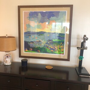 May include: A framed painting of a seascape with a town in the foreground. The painting is in a brown frame and is hanging on a wall above a dark brown wooden cabinet with a wine rack.