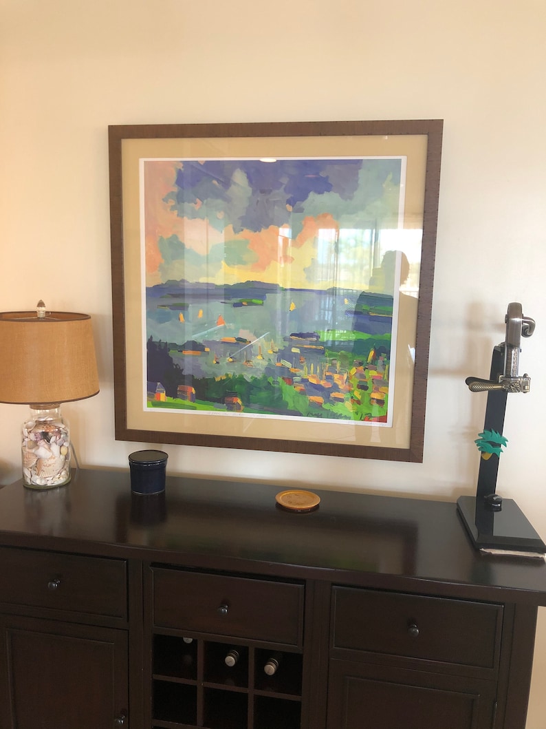 May include: A framed painting of a seascape with a town in the foreground. The painting is in a brown frame and is hanging on a wall above a dark brown wooden cabinet with a wine rack.