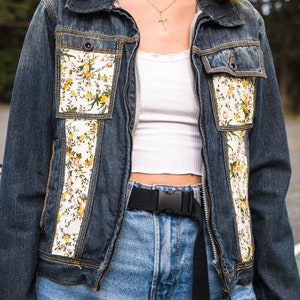 Pode incluir: Casaco de ganga azul escuro com detalhes florais e bolsos. &Eacute; usado com jeans de lavagem clara e um cinto preto. O conjunto inclui um top branco e joias douradas.