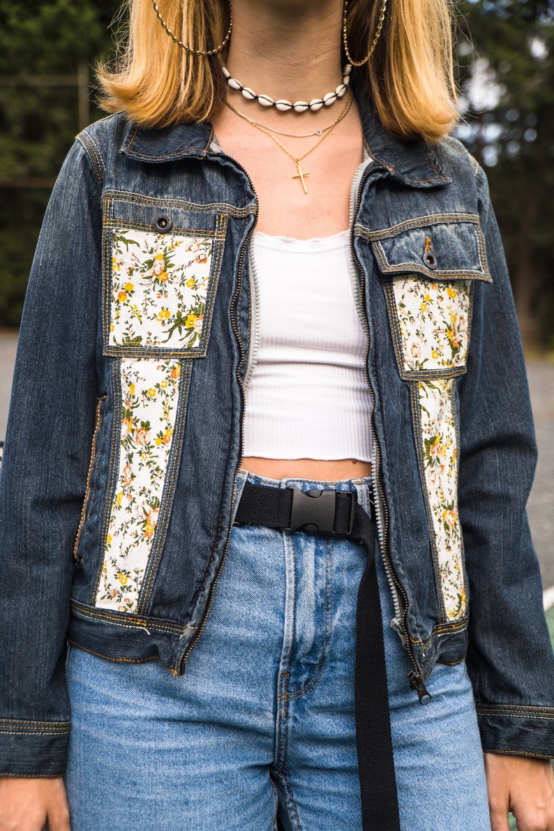Pode incluir: Casaco de ganga azul escuro com detalhes florais e bolsos. &Eacute; usado com jeans de lavagem clara e um cinto preto. O conjunto inclui um top branco e joias douradas.