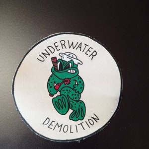 May include: A circular patch with a white background and a black border. It features a green frog wearing a white sailor hat, holding a red stick of dynamite with a lit fuse, and the text "UNDERWATER DEMOLITION" printed in black capital letters.