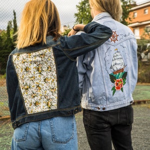 Pode incluir: Duas jaquetas jeans. Uma &eacute; azul escuro com um painel traseiro floral. A outra &eacute; azul claro com um desenho de navio e floral nas costas. Ambas as jaquetas t&ecirc;m fechos de bot&atilde;o e s&atilde;o usadas com jeans.