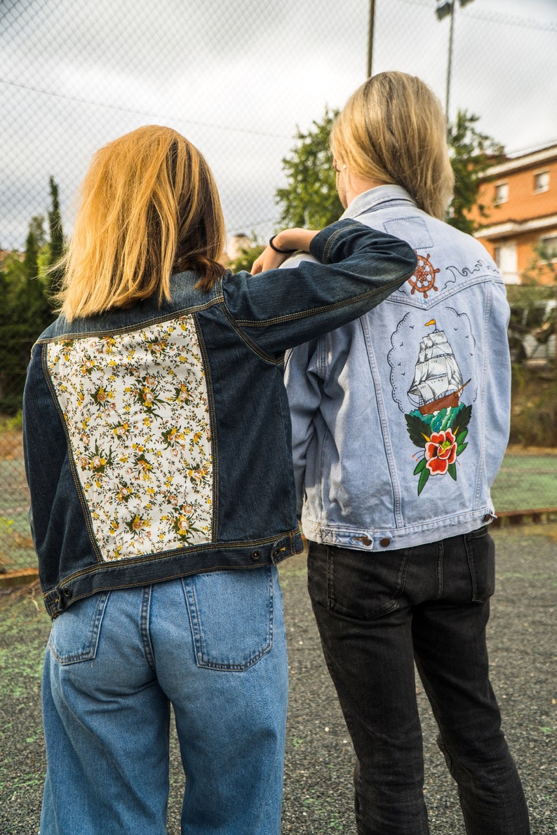 Pode incluir: Duas jaquetas jeans. Uma &eacute; azul escuro com um painel traseiro floral. A outra &eacute; azul claro com um desenho de navio e floral nas costas. Ambas as jaquetas t&ecirc;m fechos de bot&atilde;o e s&atilde;o usadas com jeans.