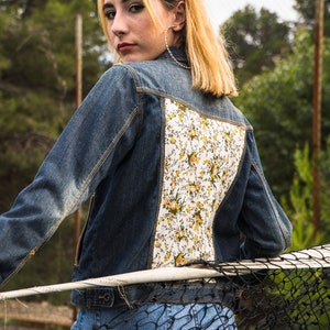 Pode incluir: Casaco de ganga azul m&eacute;dio com painel traseiro estampado floral. O padr&atilde;o floral apresenta flores amarelas e brancas sobre um fundo branco. A pessoa est&aacute; usando jeans azul.