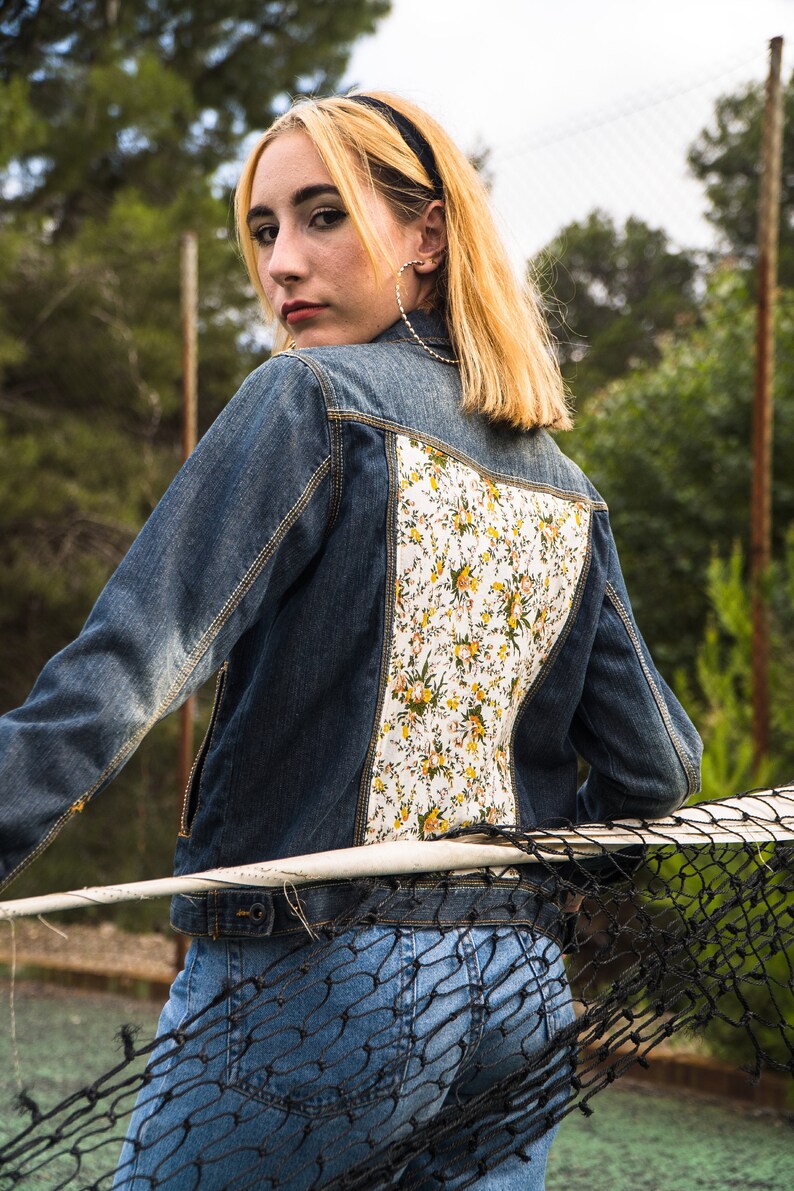 Pode incluir: Casaco de ganga azul m&eacute;dio com painel traseiro estampado floral. O padr&atilde;o floral apresenta flores amarelas e brancas sobre um fundo branco. A pessoa est&aacute; usando jeans azul.