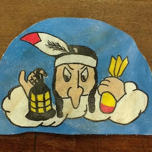 Può includere: Un cerotto di pelle dipinto a mano che raffigura un nativo americano di cartone animato con un naso lungo, che indossa un copricapo piumato, tenendo una bomba in una mano e una lanterna accesa nell'altra. Lo sfondo è blu con nuvole bianche.