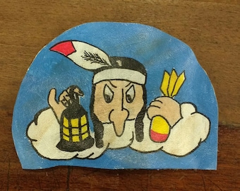Puede incluir: Un parche de cuero pintado a mano que representa a un nativo americano de dibujos animados con una nariz larga, que lleva un tocado de plumas, sosteniendo una bomba en una mano y una linterna encendida en la otra. El fondo es azul con nubes blancas.