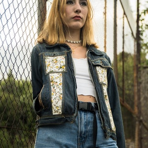 Pode incluir: Uma jaqueta jeans com detalhes florais e jeans azuis combinando. A jaqueta apresenta um padr&atilde;o floral branco nos bolsos e na frente. A modelo est&aacute; usando um top curto branco e um cinto preto. O fundo &eacute; uma cerca de tela de arame.