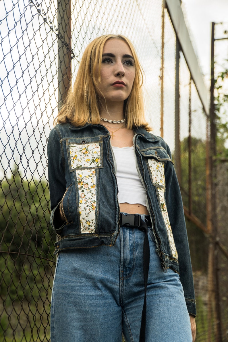 Pode incluir: Uma jaqueta jeans com detalhes florais e jeans azuis combinando. A jaqueta apresenta um padr&atilde;o floral branco nos bolsos e na frente. A modelo est&aacute; usando um top curto branco e um cinto preto. O fundo &eacute; uma cerca de tela de arame.