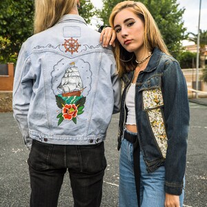 Pode incluir: Duas jaquetas jeans. Uma azul clara com um desenho de tatuagem de navio e rosa, a outra azul escura com patches florais. O desenho da jaqueta azul clara apresenta um navio marrom, uma rosa vermelha e um leme.