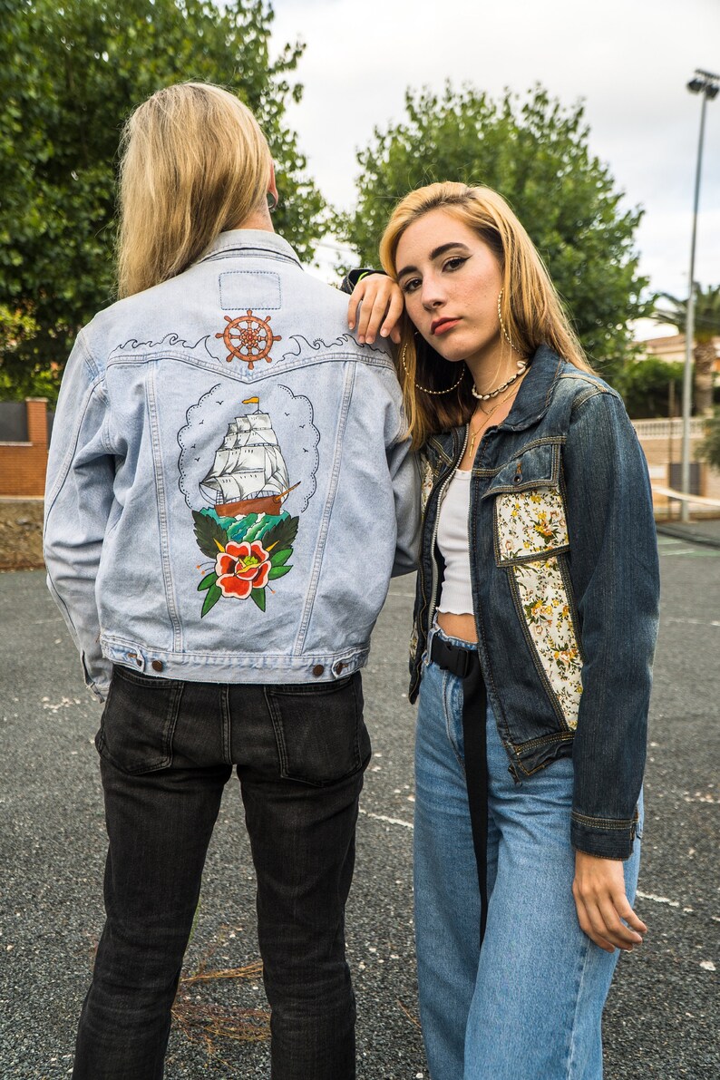 Pode incluir: Duas jaquetas jeans. Uma azul clara com um desenho de tatuagem de navio e rosa, a outra azul escura com patches florais. O desenho da jaqueta azul clara apresenta um navio marrom, uma rosa vermelha e um leme.
