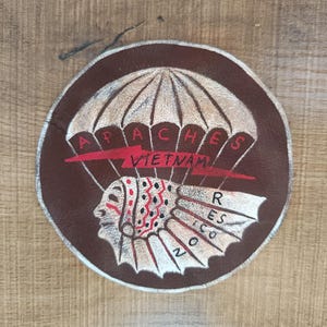 May include: A circular brown patch featuring the words "APACHES VIETNAM" and a parachute design. Below the parachute is a Native American profile with a feathered headdress. The words "RESCO" and "NO" are also visible.