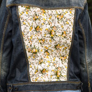 Pode incluir: Casaco de ganga azul escuro com painel floral nas costas. O padr&atilde;o floral apresenta flores amarelas, laranja e verdes sobre um fundo branco. O casaco tem fecho de bot&atilde;o e costuras amarelas.