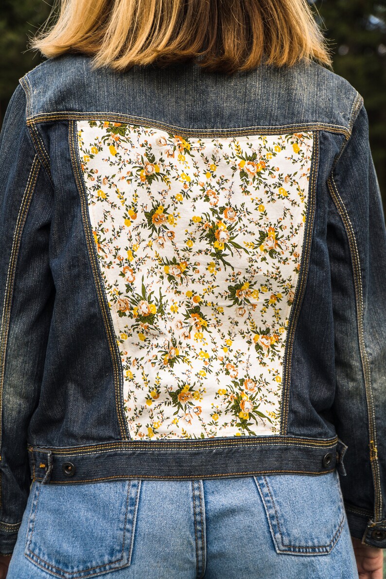 Pode incluir: Casaco de ganga azul escuro com painel floral nas costas. O padr&atilde;o floral apresenta flores amarelas, laranja e verdes sobre um fundo branco. O casaco tem fecho de bot&atilde;o e costuras amarelas.