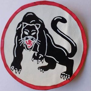 Puede incluir: Parche circular con un diseño de pantera negra en posición amenazante, con la boca roja abierta y detalles blancos. La pantera se encuentra sobre un fondo crema, enmarcada por un borde rojo. Diseño gráfico y llamativo.