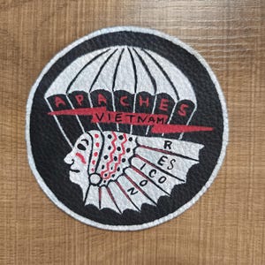 Puede incluir: Parche redondo negro con diseño de paracaídas blanco y texto "APACHES VIETNAM". Presenta un perfil nativo americano con detalles rojos y blancos. El parche es probablemente un emblema o insignia.