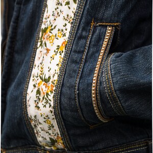 Pode incluir: Close-up de uma jaqueta jeans azul escuro com detalhes florais. A jaqueta apresenta um painel de tecido branco na frente e no bolso, adornado com flores amarelas e laranja e folhas verdes. Costuras douradas e z&iacute;per.