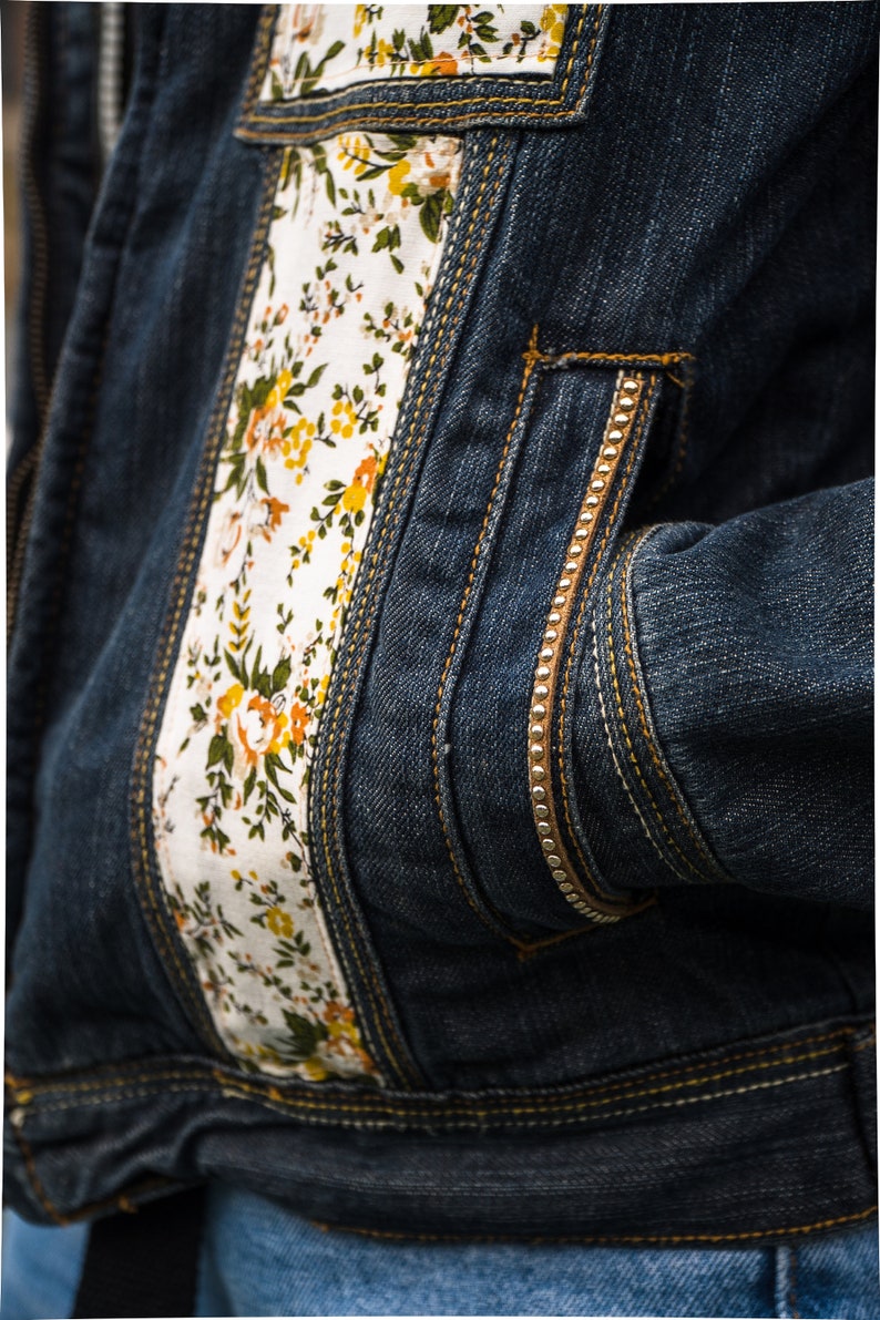 Pode incluir: Close-up de uma jaqueta jeans azul escuro com detalhes florais. A jaqueta apresenta um painel de tecido branco na frente e no bolso, adornado com flores amarelas e laranja e folhas verdes. Costuras douradas e z&iacute;per.